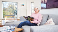 Ältere Dame sitzt lesend auf dem Sofa. Um den Hals trägt sie ein Hausnotrufgerät. Foto: Malteser
